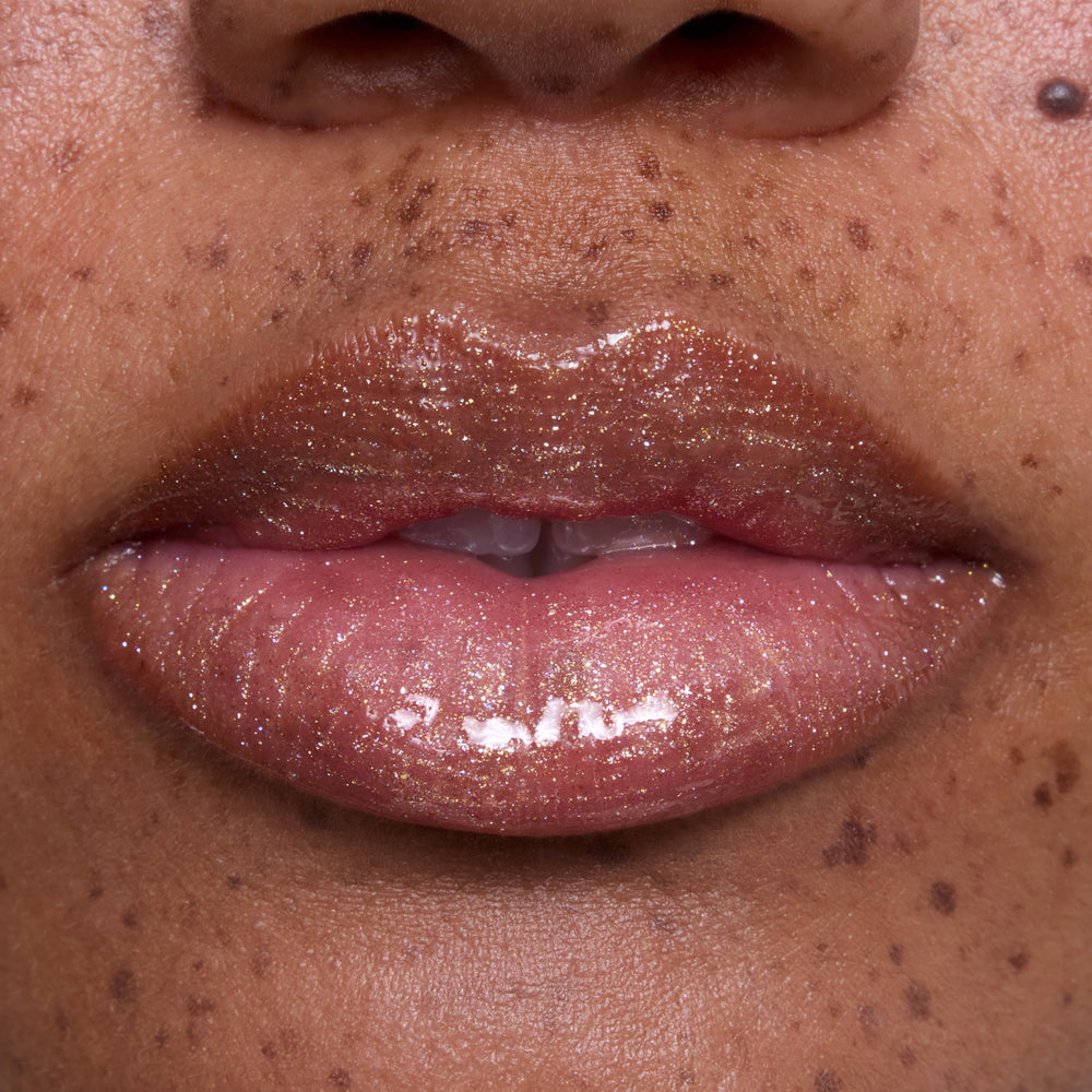 Close-up of glossy, medium-toned lips wearing Ambitious Tiana Ultra Glossy Lip, showing shimmering gold and pink sparkle inspired by Disney Princess beauty, with natural skin and freckles softly visible around the mouth.