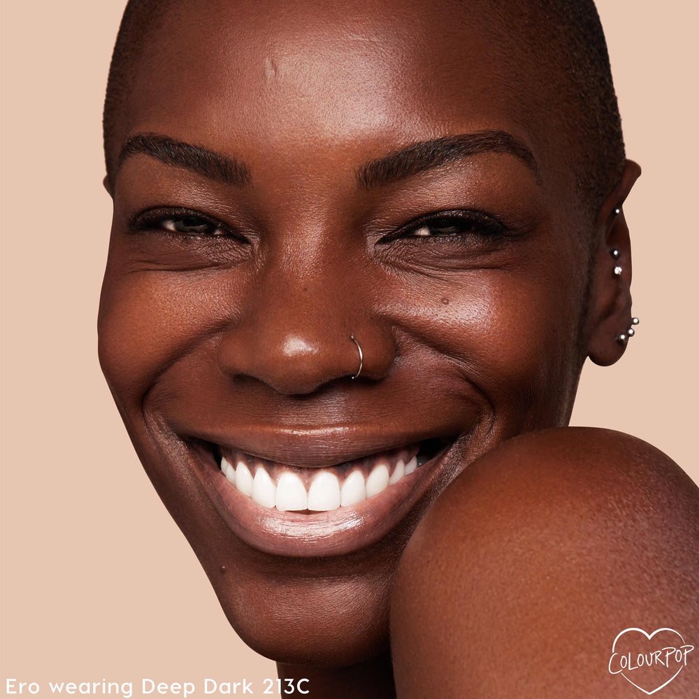 Smiling model with deep brown skin wears ColourPop Hyaluronic Hydrating Foundation in Deep Dark 213 C, showing smooth, dewy, even coverage; close-up face with nose ring and ear piercings against a warm beige background.