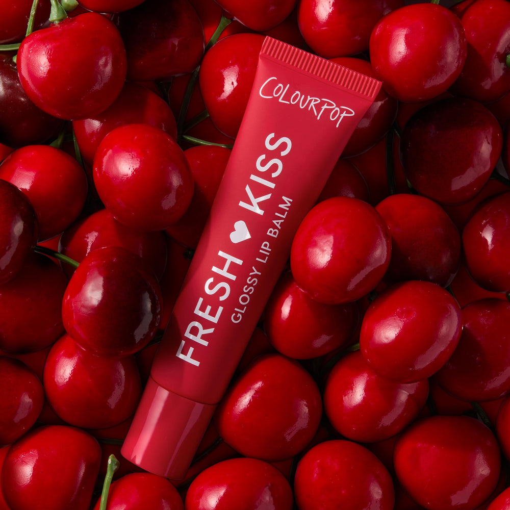 ColourPop Fresh Kiss Glossy Lip Balm in Cherry Glaze rests among glossy red cherries in a top-down flatlay, showcasing the Fruit Basket Glossy Lip Balm Trio - ColourPop Glossy Lip Balm Trio offering hydrating, high-shine cherry tint.