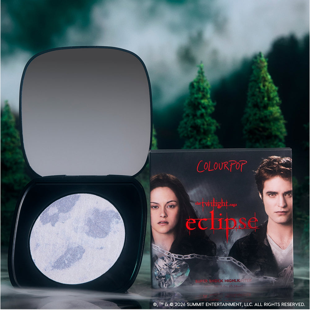 Change Me mega highlighter compact by ColourPop opened to show a silver moon-patterned pan beside its Twilight Eclipse-themed box featuring main characters, styled outdoors against a misty pine forest background.